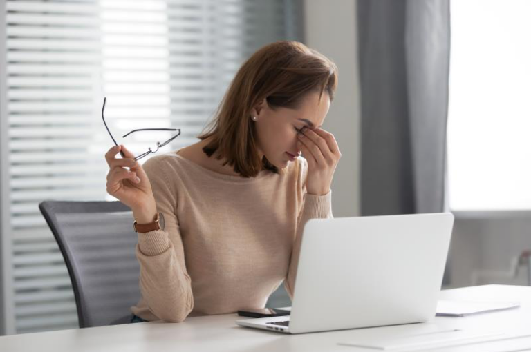 kobieta siedząca przy laptopie, ściągnęła okulary, jedną ręką trzyma się u nasady nosa