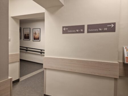 Zdjęcie przedstawia korytarz na pierwszym piętrze w okolicy pochylni wewnętrznej. Na ścianie tabliczki wskazujące numery gabinetów oraz plakaty. Po obu stronach pochylni znajdują się pochwyty.