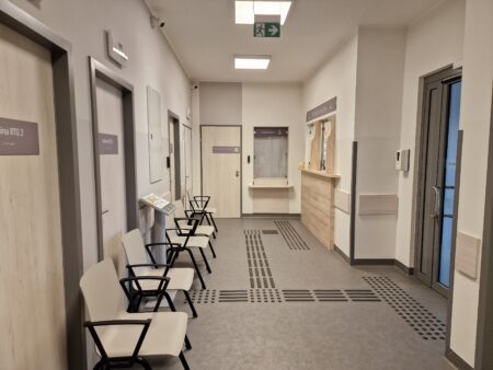 Zdjęcie przedstawia korytarz przy rejestracji głównej. Od wejścia do planu tyflograficznego oraz okienka rejestracji prowadzą linie naprowadzające. Przy ścianie znajdują się krzesła z podłokietnikami.