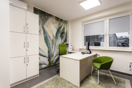 Zdjęcie przedstawia gabinet lekarski nr 1. Po lewej stronie znajduje się biurko lekarskie. Przed nim jest stołek dla pacjenta. Meble są w kolorze białym, krzesła - zielonym.