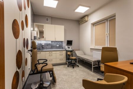 Zdjęcie przedstawia gabinet nr 13. Po lewej stronie rpzy ścianie znajduje się fotel laryngologiczny. Po prawej stronie biurko lekarskie wraz z krzesłami. W głębi widać leżankę lekarską oraz aparat USG.