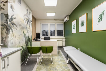 Zdjęcie przedstawia gabinet nr 4. Po prawej stronie znajduje się leżanka podnoszona elektrycznie, na wprost wejścia jest biurko lekarskie a przed nim dwa krzesła dla pacjentów.