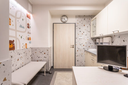 Zdjęcie przedstawia gabinet zabiegowy dziecka chorego. Po lewej stronie znajduje się leżanka dla pacjentów. Po prawej - biurko. Na wprost drzwi a nad nimi zegar.