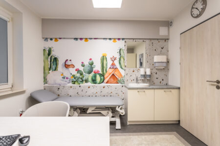 Zdjęcie przedstawia gabinet lekarski dziecka chorego. Na wprost widoczna jest leżanka lekarska, nad nią na ścianie znajduje się tapeta z lamą. Zdjęcie robione jest zza biurka lekarskiego, przed którym stoi krzesło dla pacjenta.