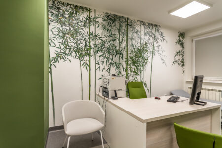 Zdjęcie przedstawia gabinet lekarski nr 3. Po lewej stronie od wejścia przed ścianą z tapetą w bambusy stoi biurko lekarskie. Przed nim znajdują się krzesła dla pacjentów.