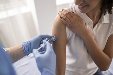 Obrazek przedstawia zabieg szczepienia - osoba w niebieskich rękawiczkach wstrzykuje młodej kobiecie preparat